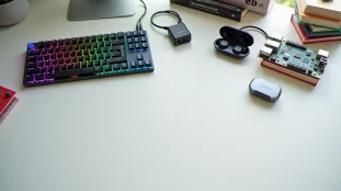 Sélection soignée de cadeaux tech disposés sur un bureau blanc en bois, illustrant un tech guide gift site avec clavier mécanique, écouteurs sans fil, hub USB-C et livres techniques de programmation